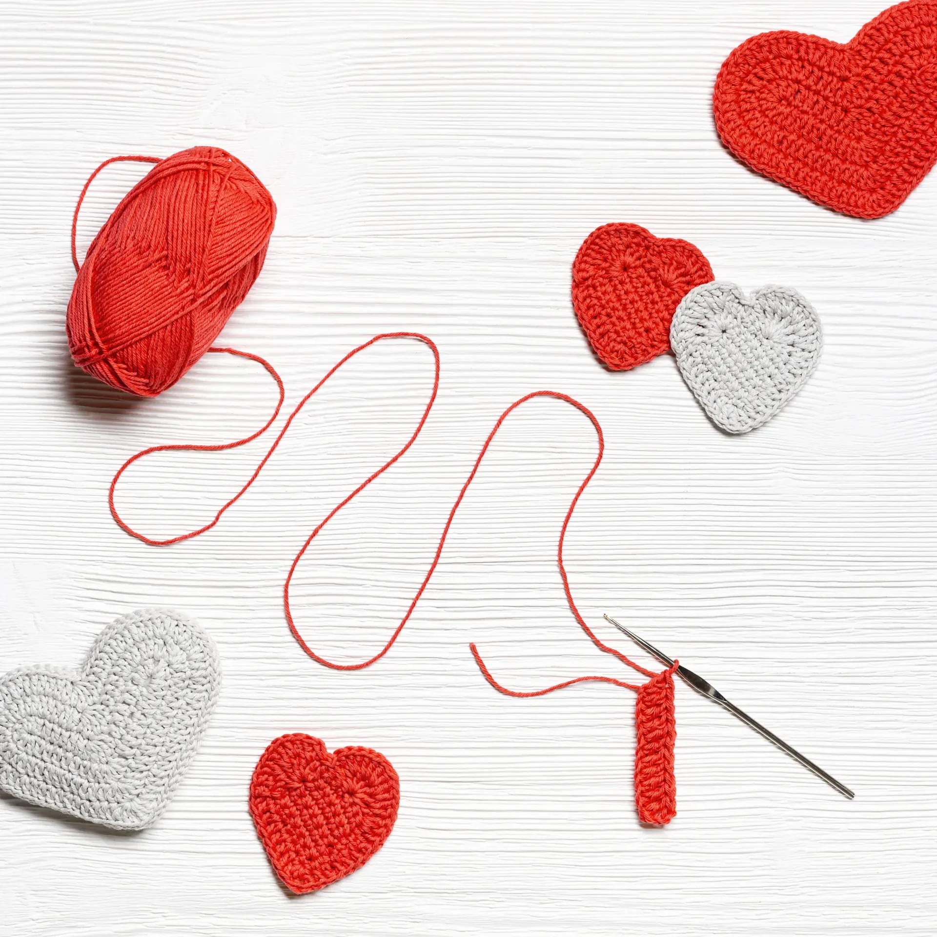 Red and grey crocheted hearts, a ball of red yarn and a crochet hook on a white wooden surface